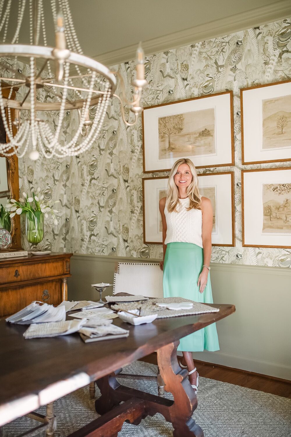 Alex Hechart standing in a beautifully decorated room with a large wooden table, white chandelier, and a green patterned wallpaper with gold picture frames.