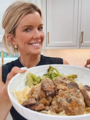 Jamie Tarence of Family Savvy holding a bowl of homemade Swedish meatballs with creamy mushroom sauce, served over egg noodles with a side of broccoli