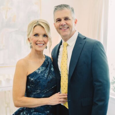 Portrait shot of Jamie Tarence wearing a one-shoulder dark blue floral dress with her husband Zane Tarence wearing a navy suit with a yellow tie