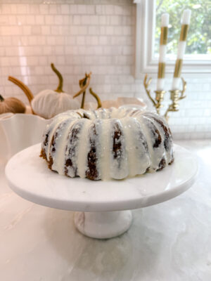 Family Savvy's Pumpkin Carrot Bundt Cake with Cream Cheese Frosting on a white marble cake pedestal on a kitchen counter 