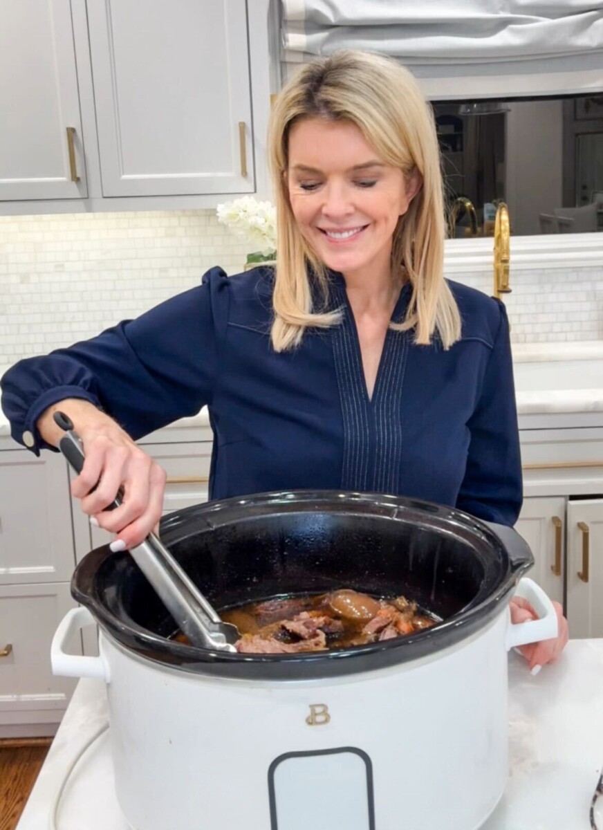 Jamie Tarence of Family Savvy standing in her kitchen looking down at a slow cooker filled with a rump roast and vegetables