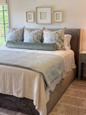 Close-up of a made bed in one of Jamie's lake house bedrooms. The bed is made with a linen bed cover along with a variety of green pillows and blankets.