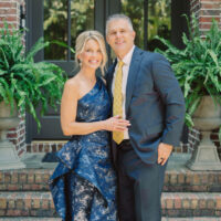 Jamie Tarence dressed in a floor-length blue gown standing next to her husband Zane Tarence dressed in a suit outside their home