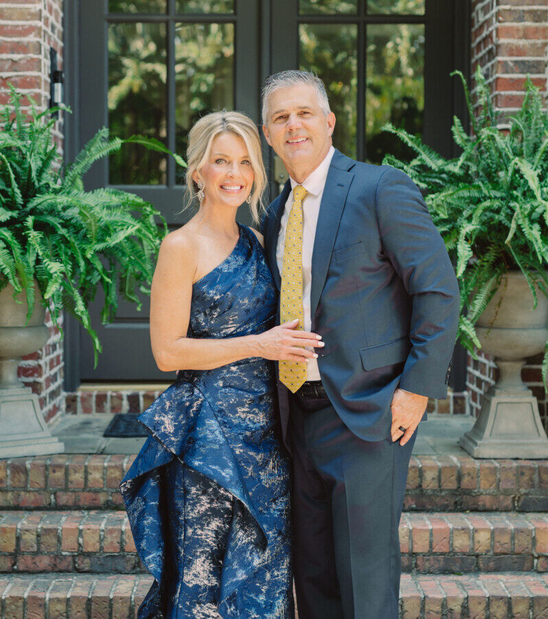 Jamie Tarence dressed in a floor-length blue gown standing next to her husband Zane Tarence dressed in a suit outside their home